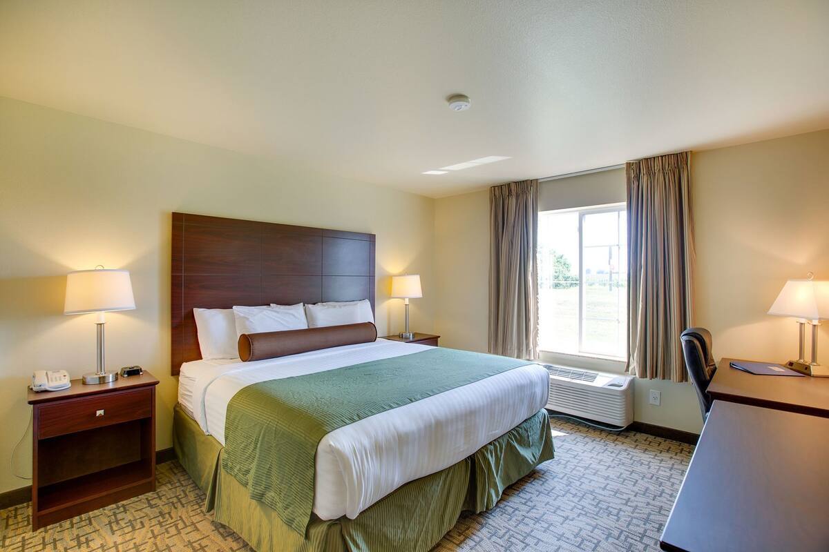 A hotel room with a bed, two bedside tables with lamps, a desk with a chair, a window with curtains, and a radiator.