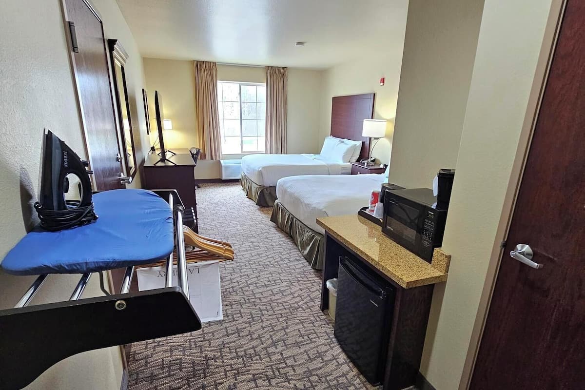 A hotel room with two beds, a desk with a computer, a chair, a small kitchenette, and a window with curtains.