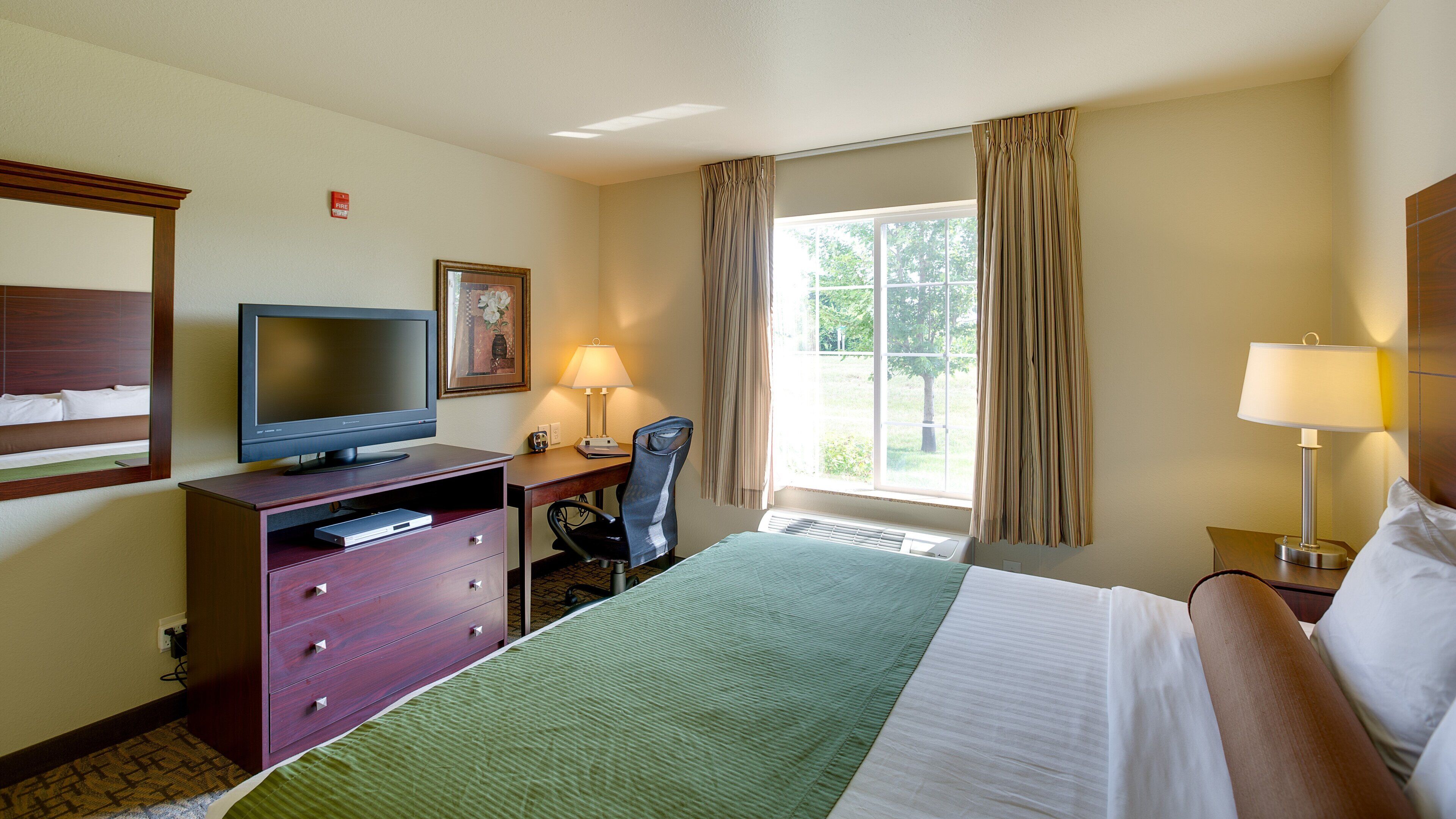 A hotel room with a bed, a desk with a computer, a television, a lamp, and a window with curtains.