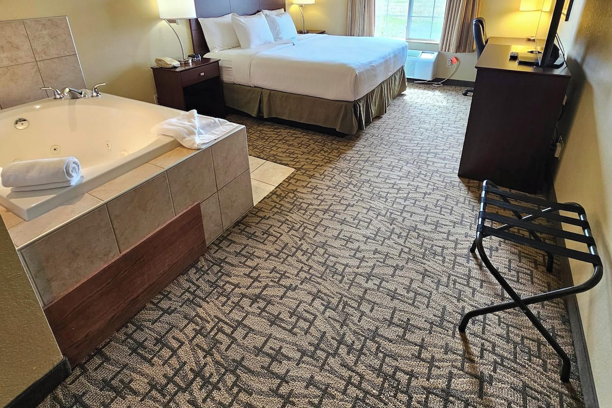 A hotel room with a large bed, a separate bathroom with a bathtub, a desk with a computer, and a window with curtains.
