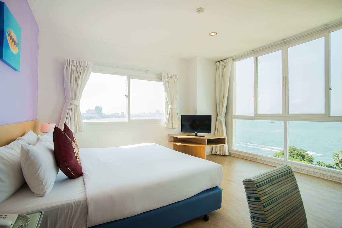 A hotel room with a large bed, a TV on a wooden stand, and a view of the ocean.