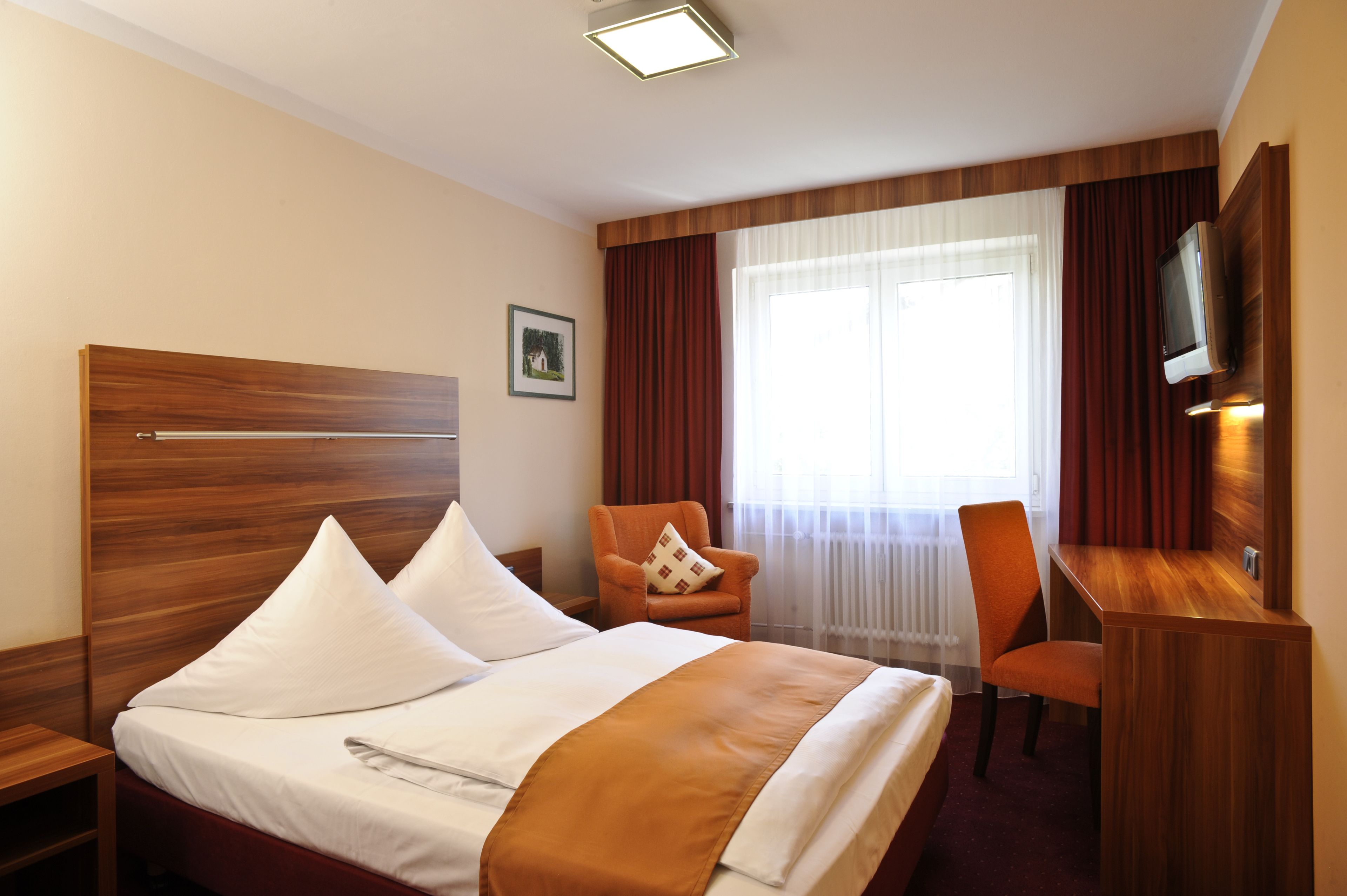 Habitación de hotel con una cama de cabecera de madera, un escritorio con silla, televisión y ventana con cortinas.