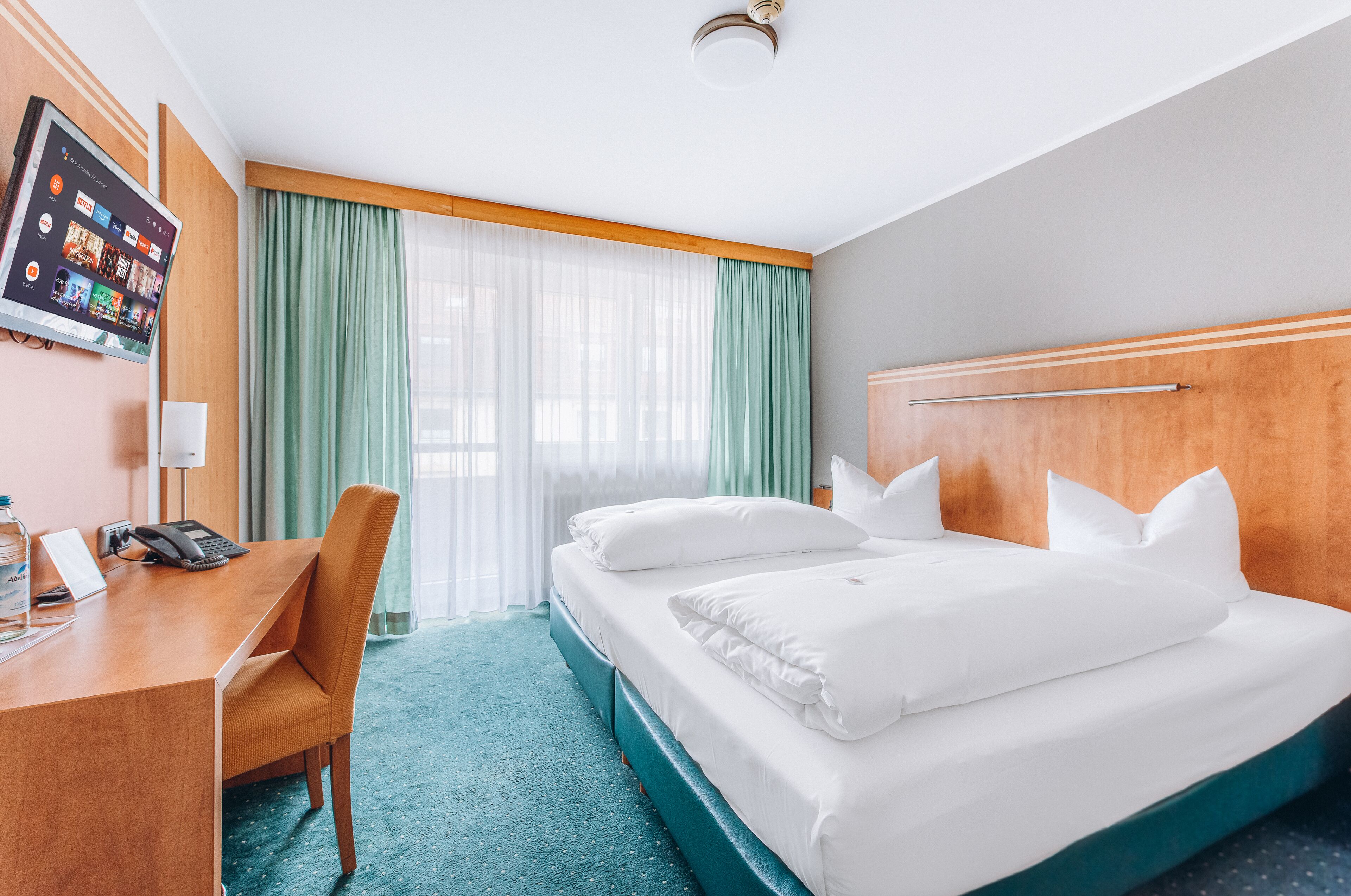 Una habitación de hotel con cama, escritorio, teléfono, botella, televisor en la pared y ventana con cortinas.