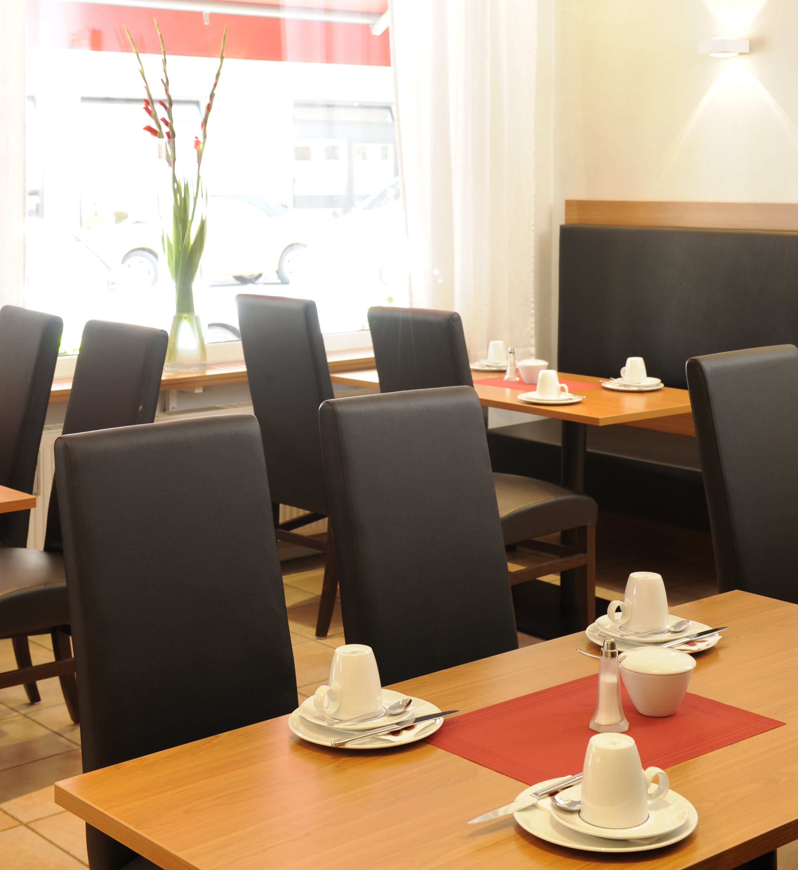 Un comedor con mesas dispuestas para dos, sillas negras y un jarrón con flores en el alféizar.