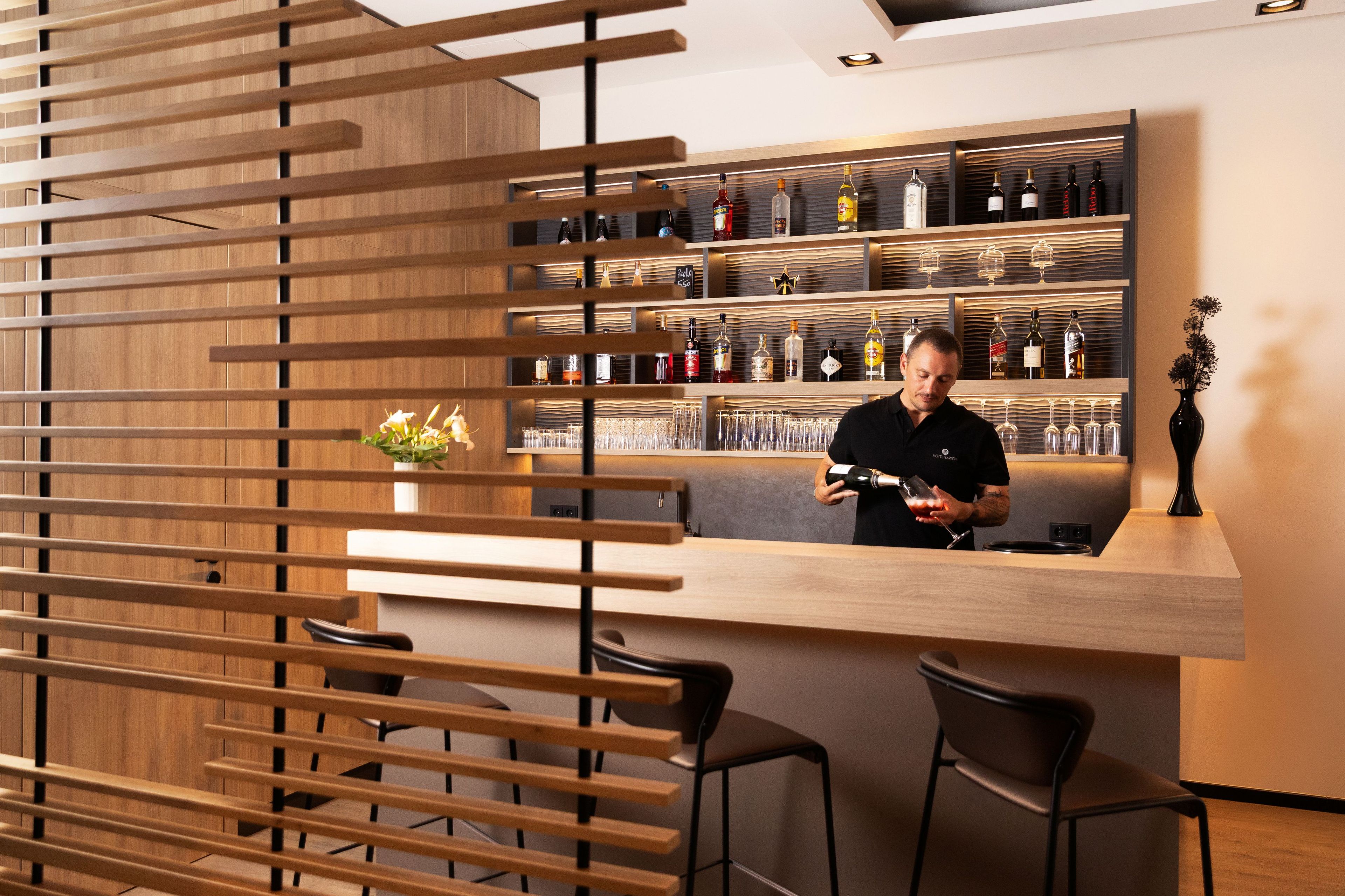 A bar area with a wooden slatted partition, a counter with a person behind it, and shelves stocked with bottles and glasses.
