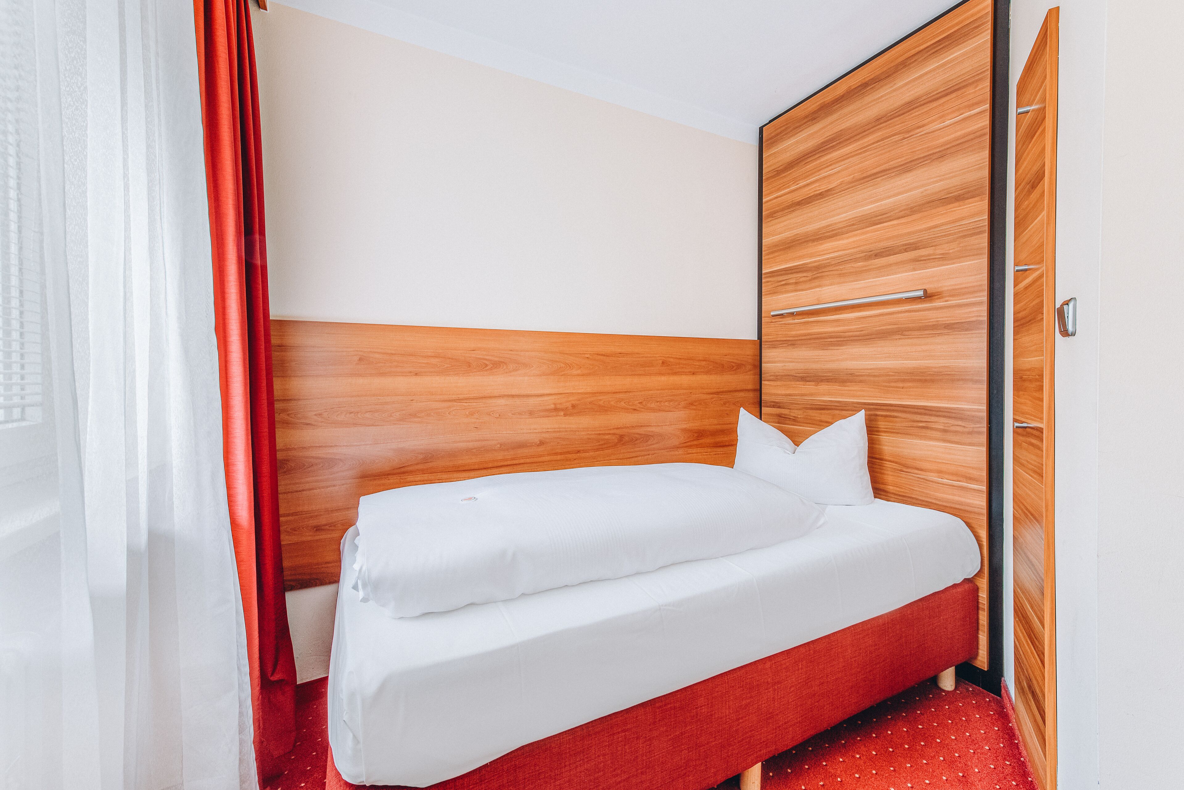 Habitación de hotel con cabecera de madera, una cama individual con ropa blanca, alfombra roja y cortina roja.