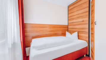 Habitación de hotel con cabecera de madera, una cama individual con ropa blanca, alfombra roja y cortina roja.