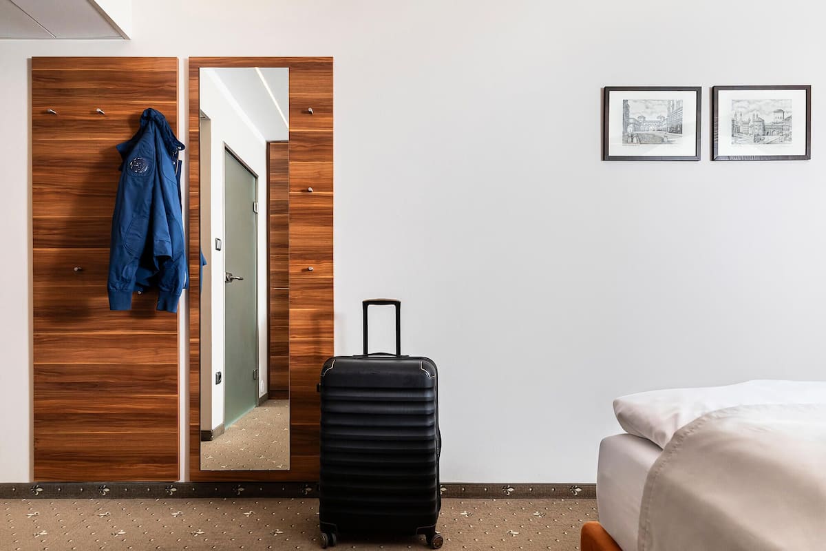 A hotel room with a wooden wardrobe, a black suitcase, a bed with white bedding, and two framed pictures on the wall.