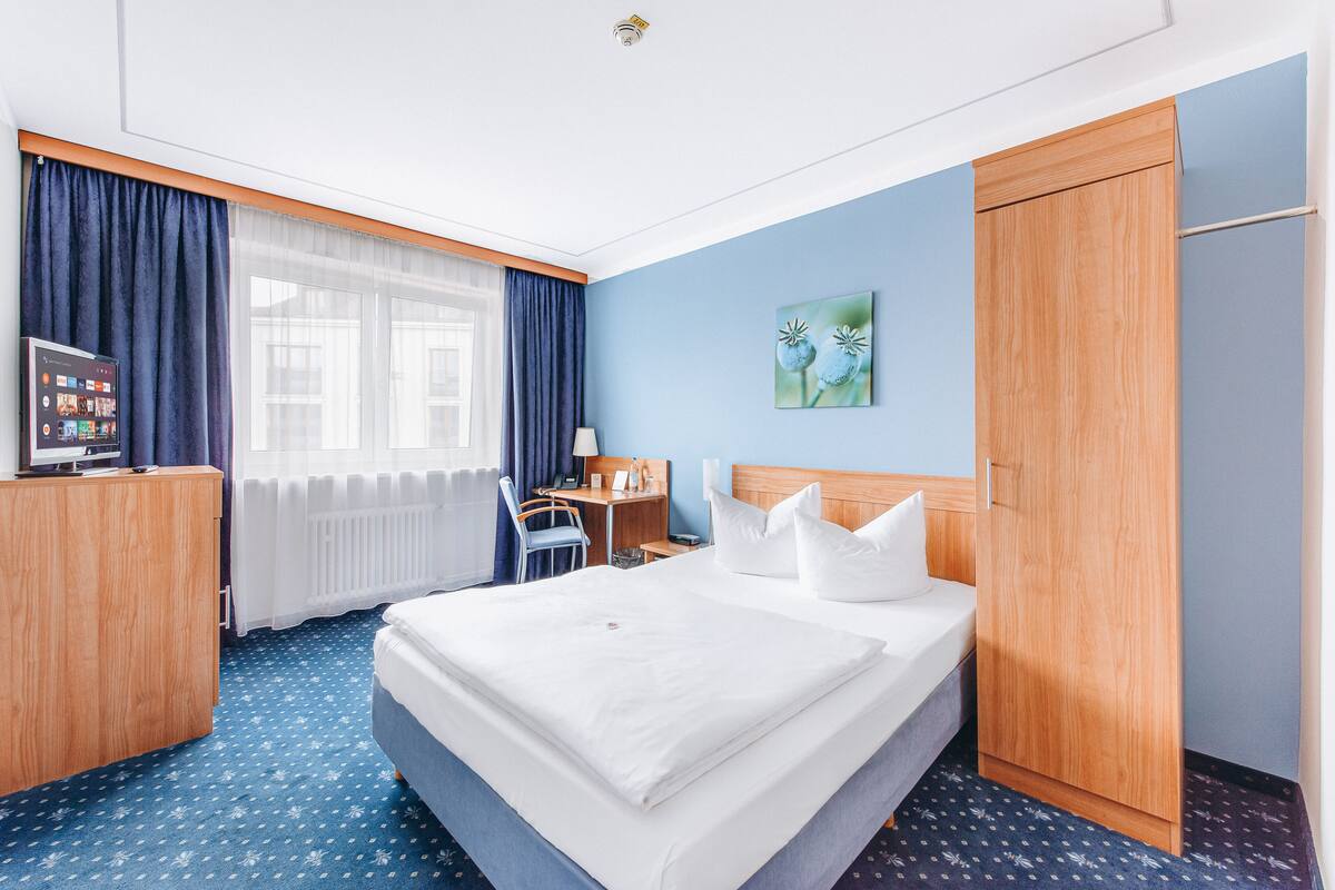 A hotel room with a large bed, a desk, a chair, a TV, and a wardrobe.