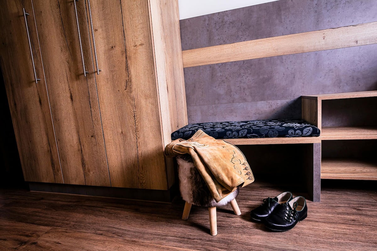 A wooden wardrobe, a bench with a patterned cushion, and a pair of shoes on a small stool.