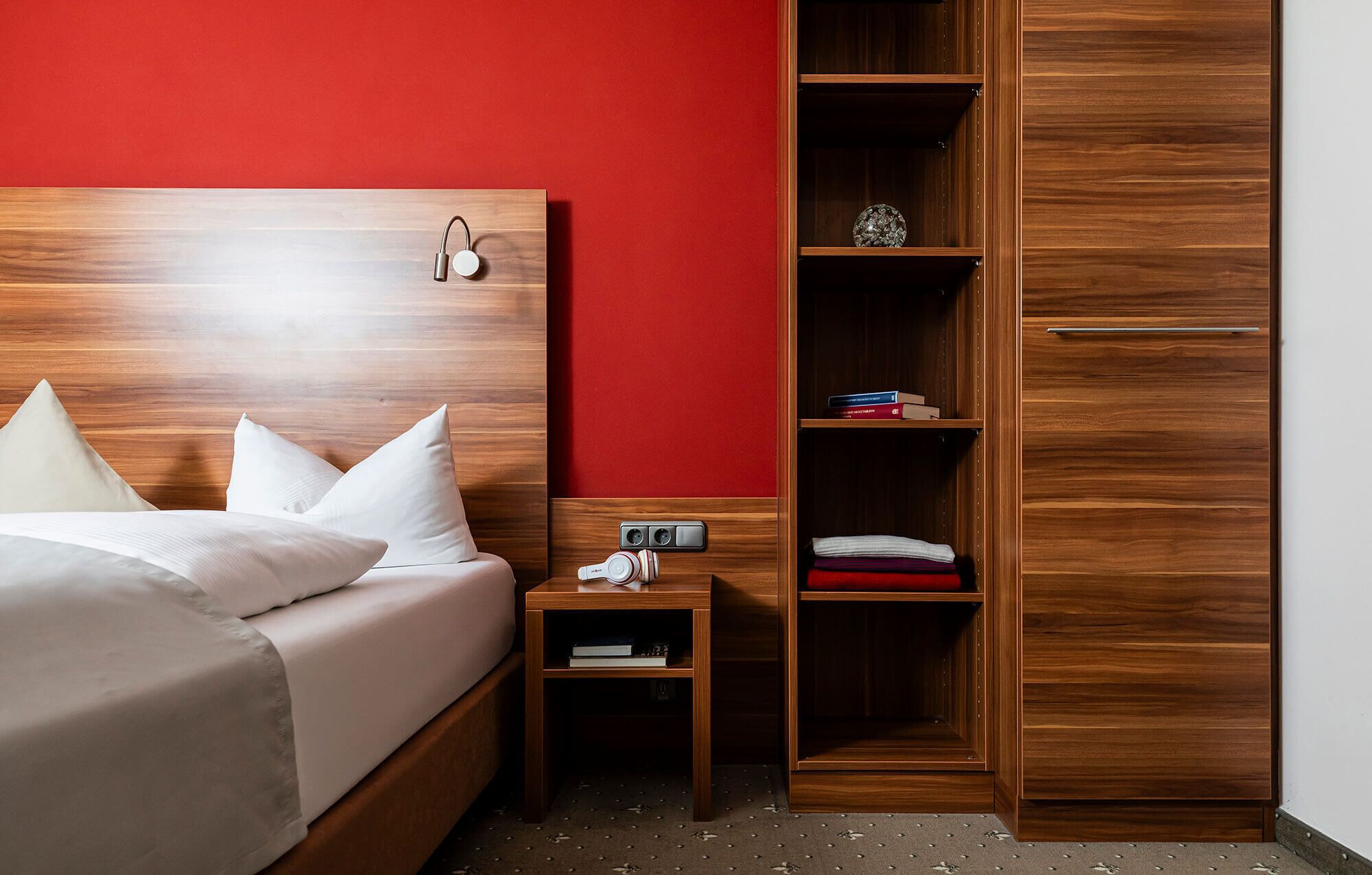 Habitación de hotel con cabecera de madera, cama con ropa blanca, mesita de noche con teléfono, pared roja y armario de madera.