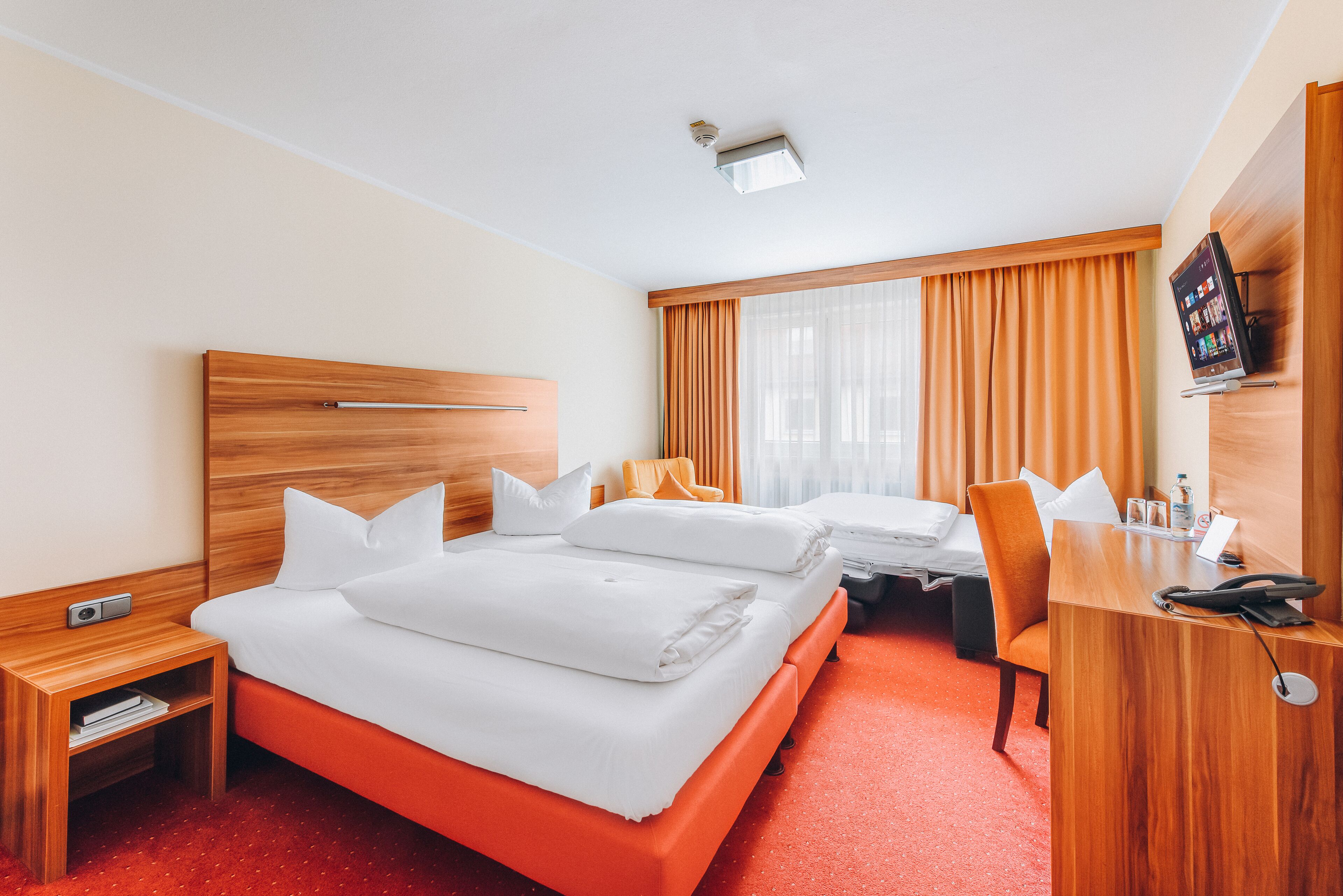 A hotel room with a wooden headboard, two beds with white bedding, a desk with a chair, a television, and a small table with a bottle and glasses.