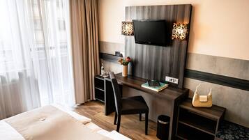Una habitación de hotel con cama, un escritorio con silla, televisor, un florero con flores, lámpara y una mesita con bolso.