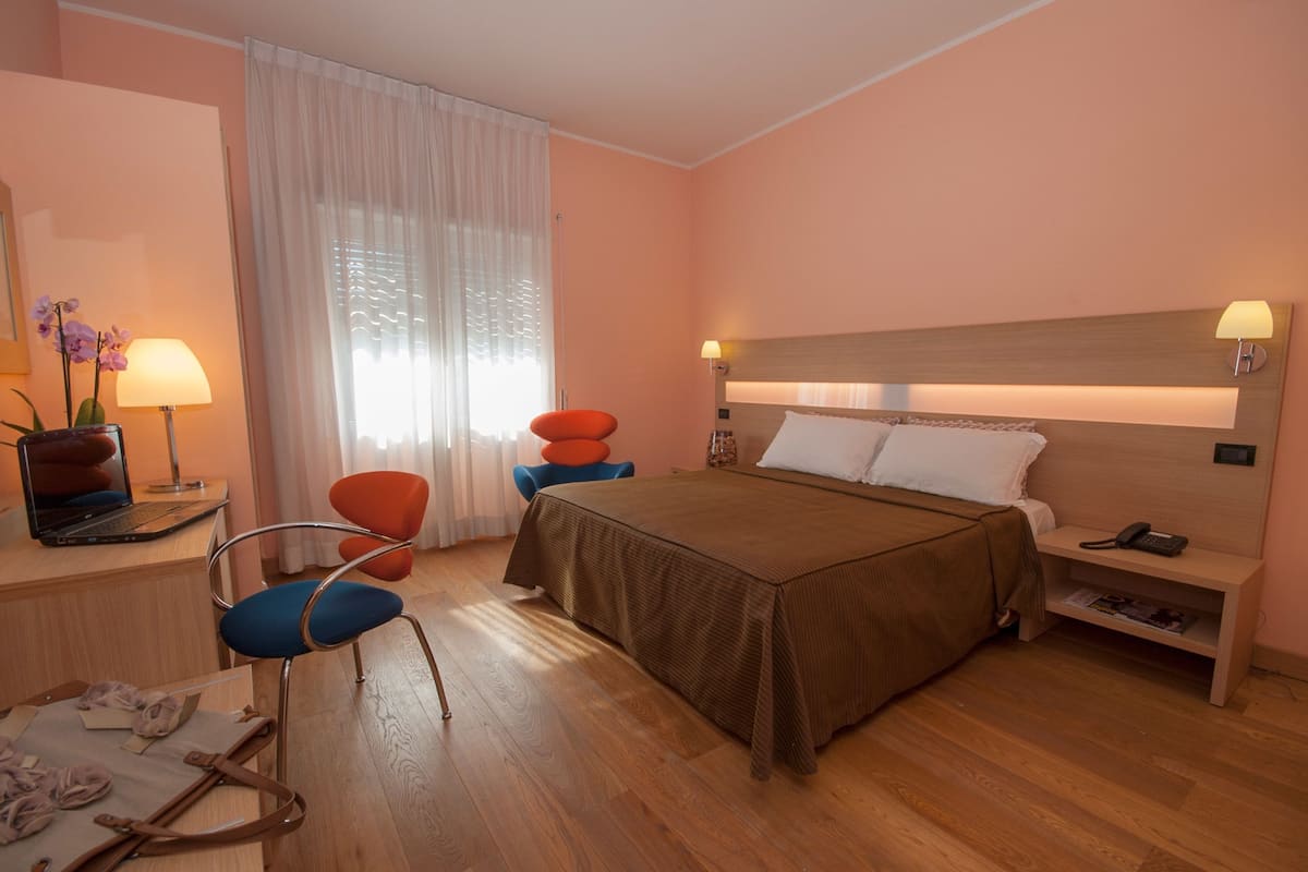 Habitación de hotel con cama, mesita de noche, lámpara, silla, escritorio y ventana con cortinas.
