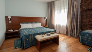 Habitación de hotel con cabecera de madera, una cama grande con ropa de cama estampada, una mesita de noche con una lámpara, una pequeña mesita de noche con un teléfono, un banco con un libro y una tableta, y una ventana con cortinas a rayas.
