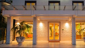 Entrada de un hotel con un corredor cubierto, plantas en macetas grandes y puerta de vidrio.