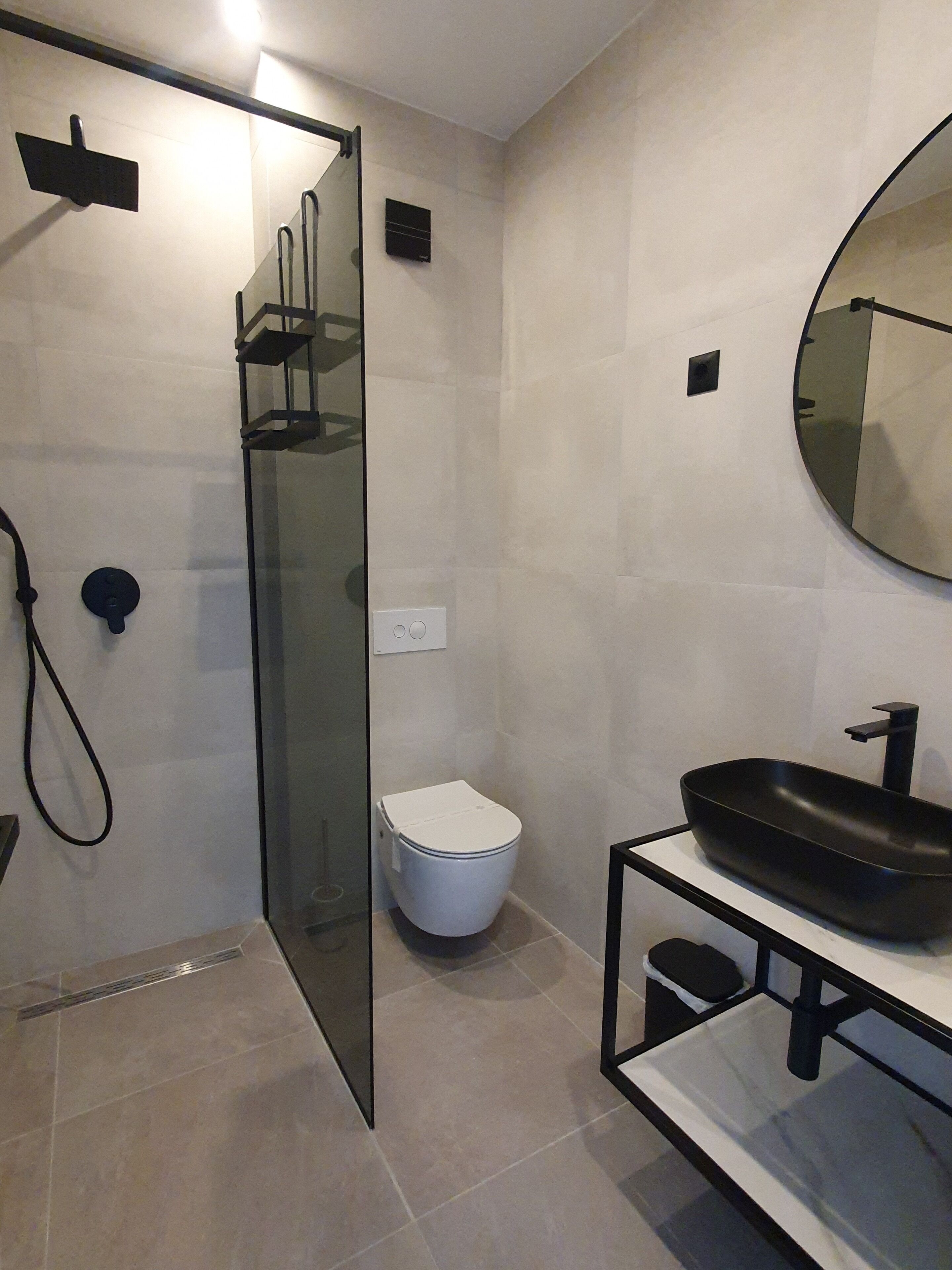 Un baño moderno con cabina de ducha de vidrio, inodoro blanco y lavamanos negro con un espejo redondo encima.