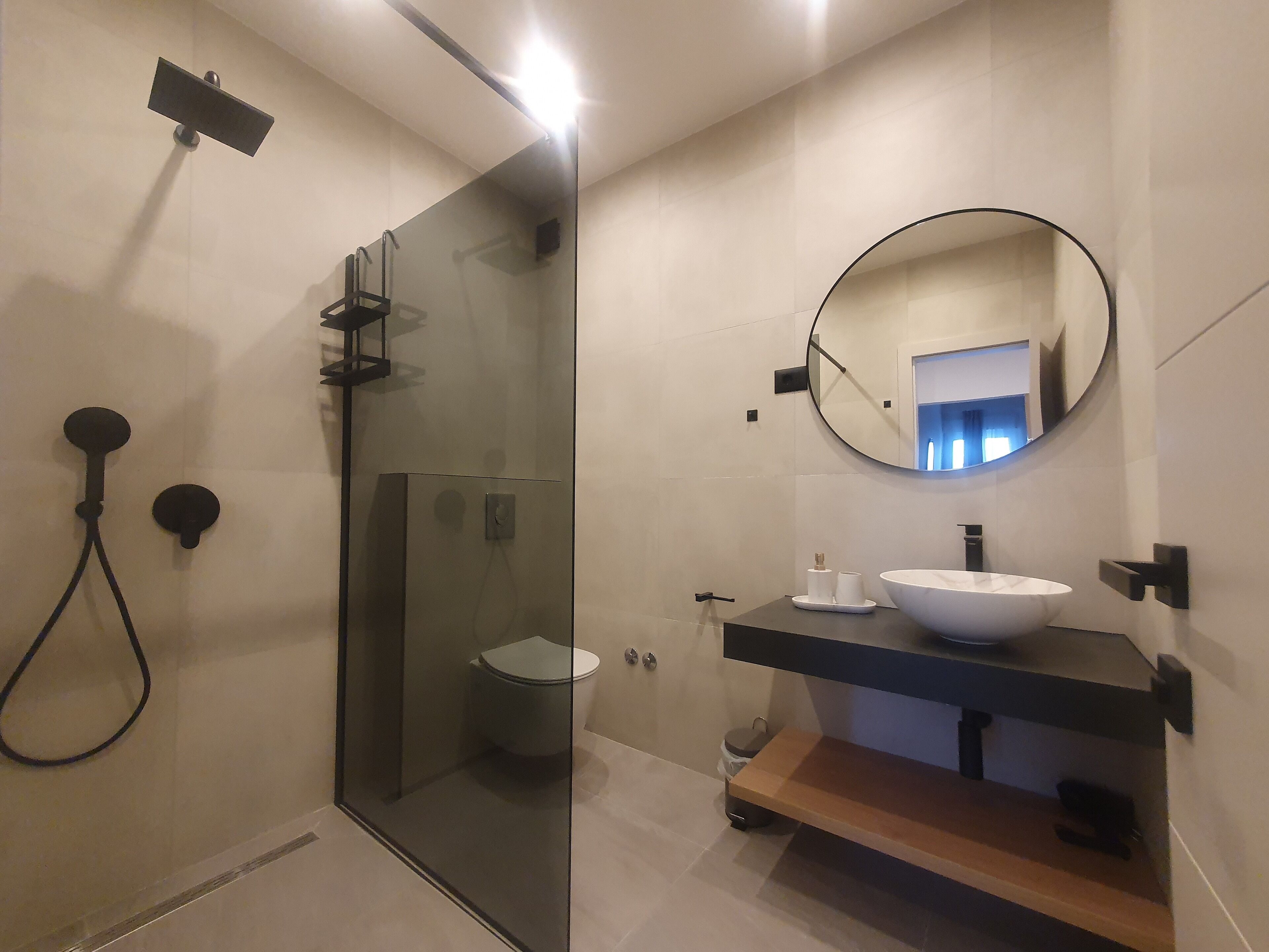 A modern bathroom with a glass shower enclosure, a round mirror, and a white sink.