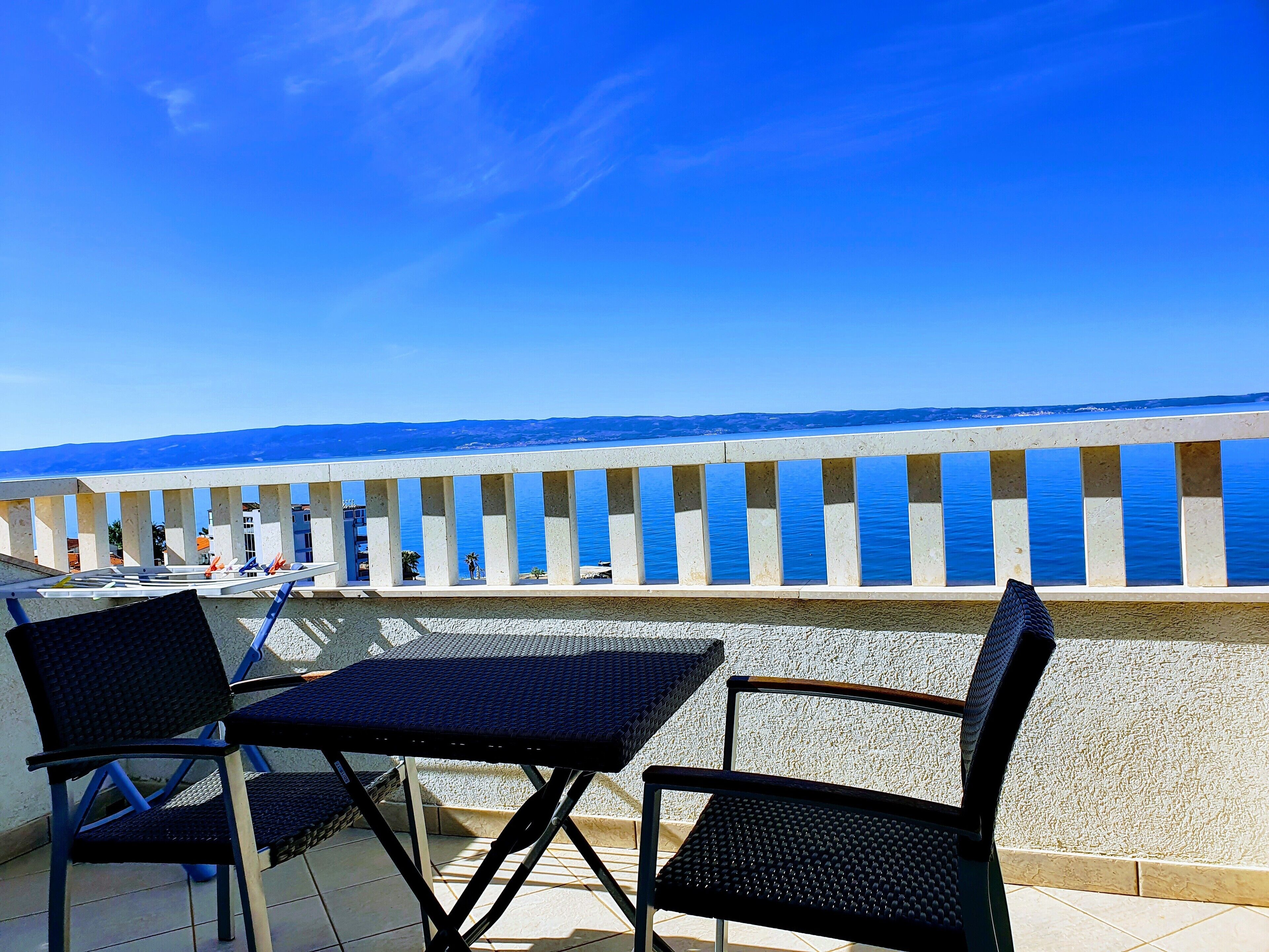Eine Terrasse mit zwei schwarzen Metallstühlen und einem Tisch, mit Blick auf ein ruhiges Meer und einen wolkenlosen, blauen Himmel.