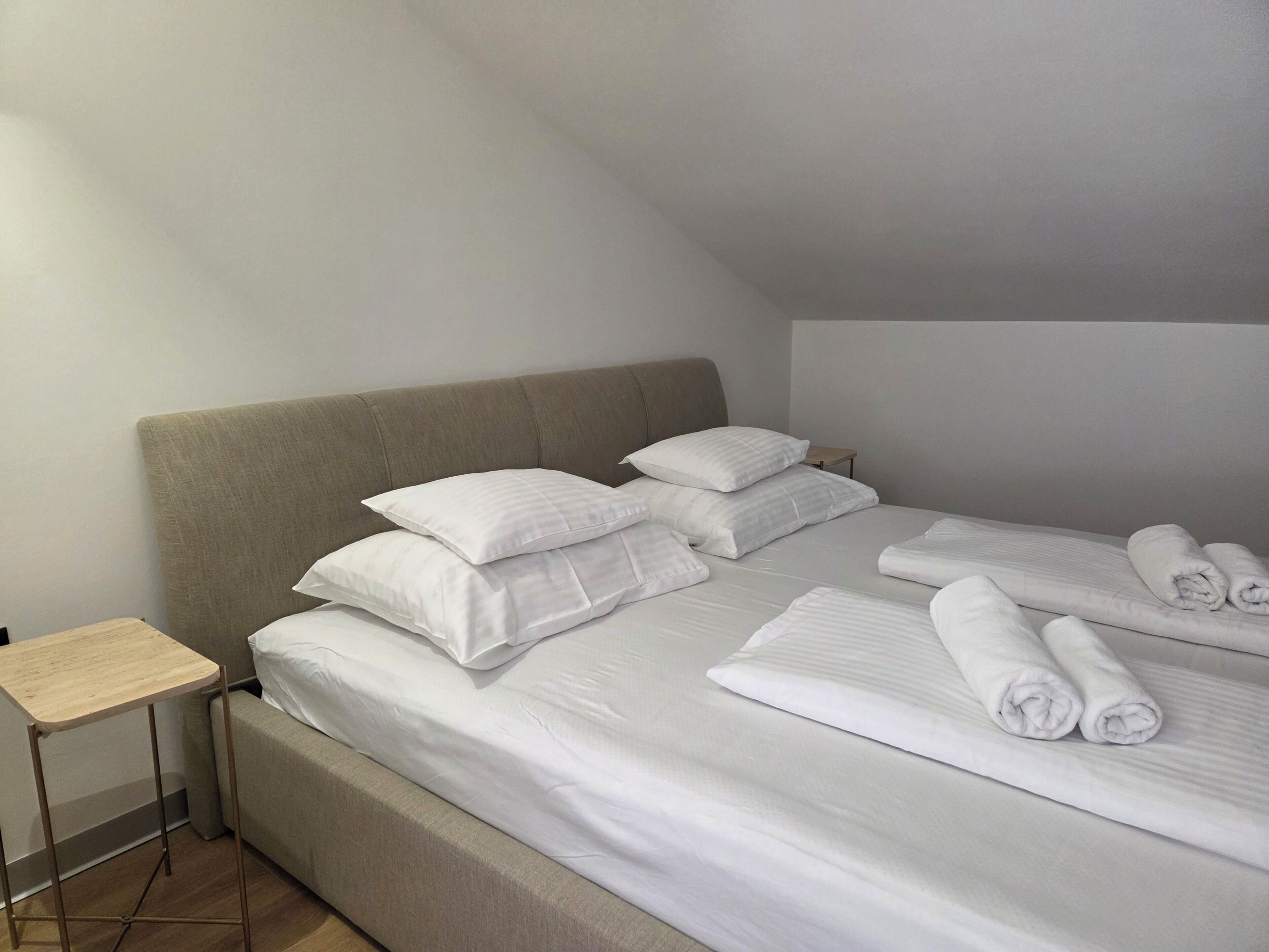 A neatly made bed with white linens and pillows, a small bedside table, and a wooden floor.