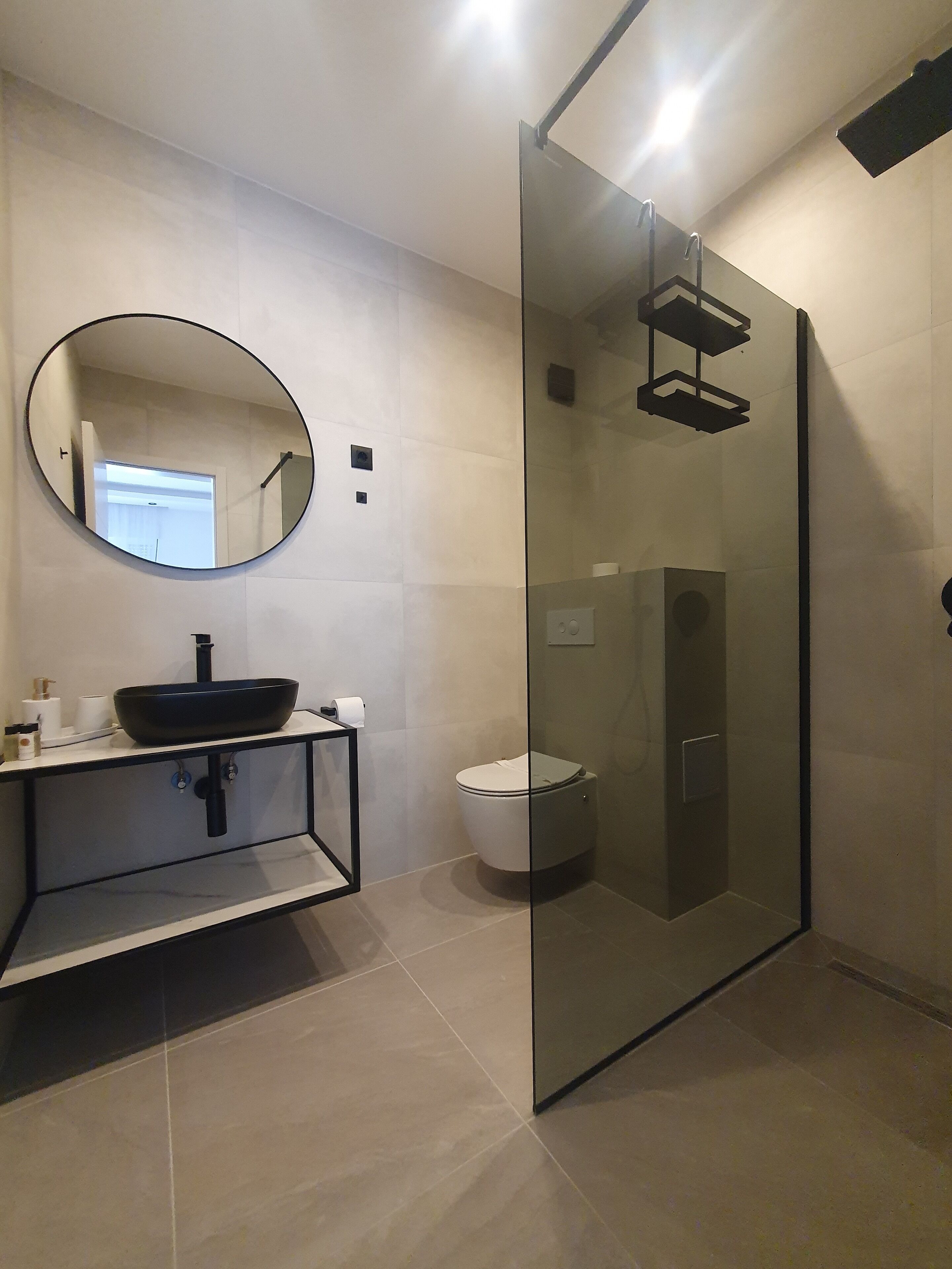 Een moderne badkamer met een douchecabine van glas, een ronde spiegel, een zwarte wastafel en een toilet.