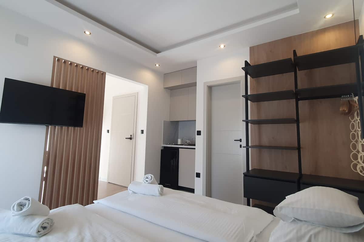 Una moderna camera d'albergo con un letto, una TV fissata al muro, un mobiletto a mensole in legno e, sullo sfondo, un angolo cottura.