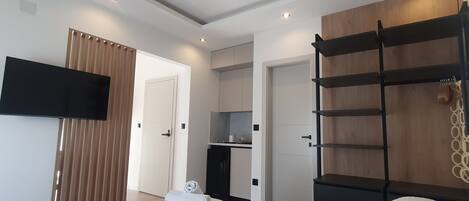 A modern hotel room with a bed, a TV mounted on the wall, a wooden shelf unit, and a kitchenette in the background.