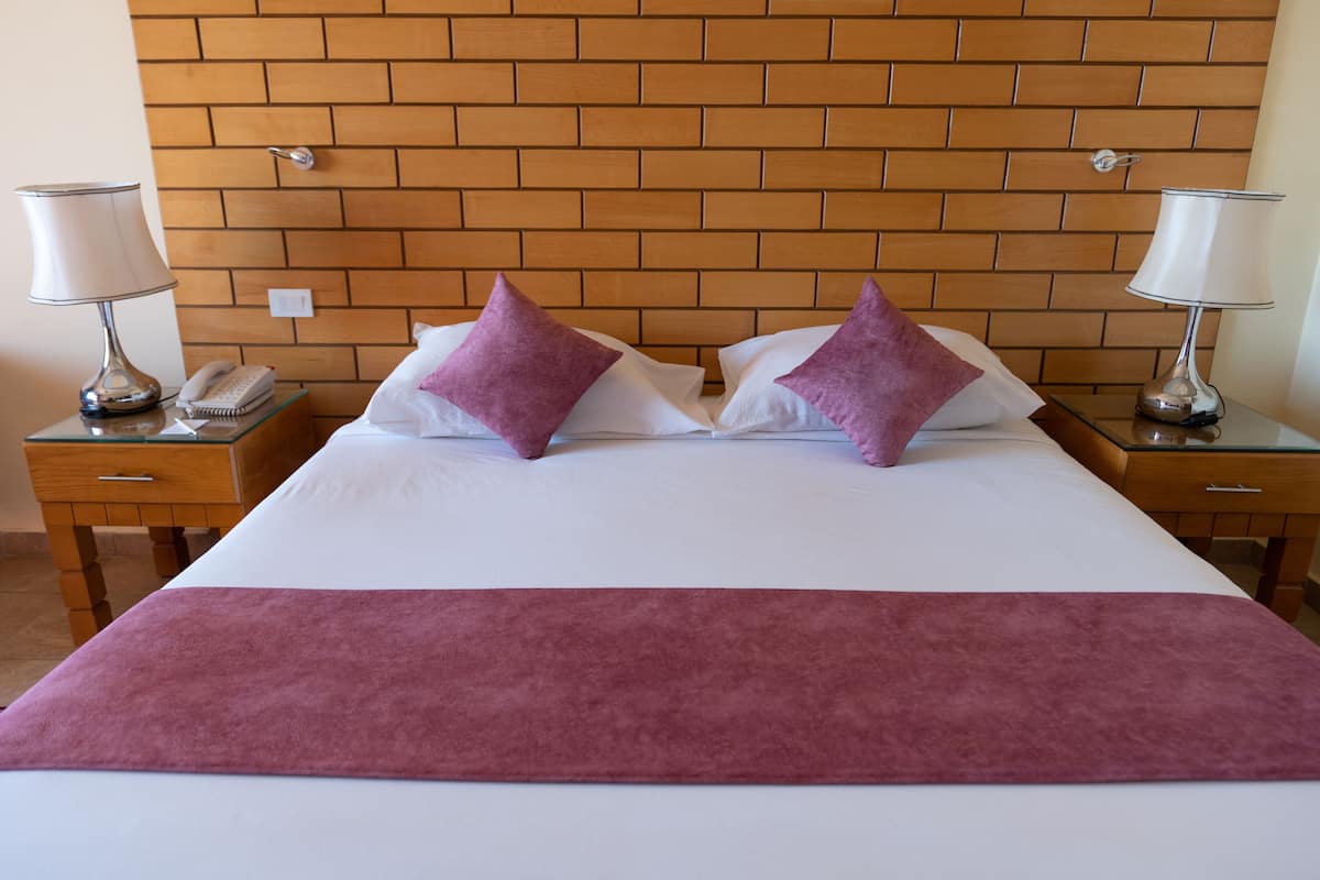 Ein Hotelzimmer mit einem Kopfende aus Backsteinoptik, zwei Nachttischen mit Lampen, einem Telefon und zwei violetten Kissen.