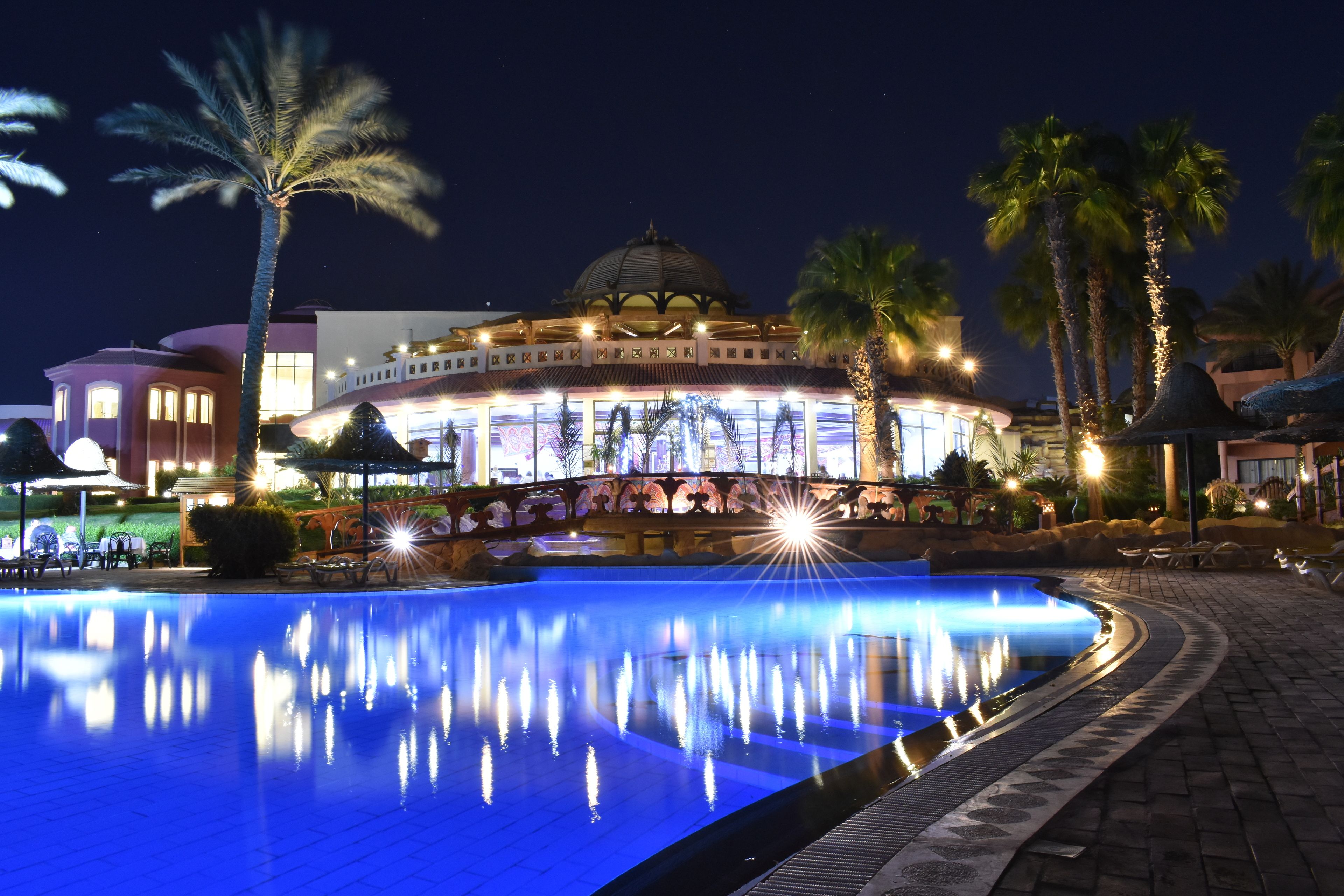 Una piscina con una fuente, rodeada de palmeras y un edificio con cúpula y arcos.