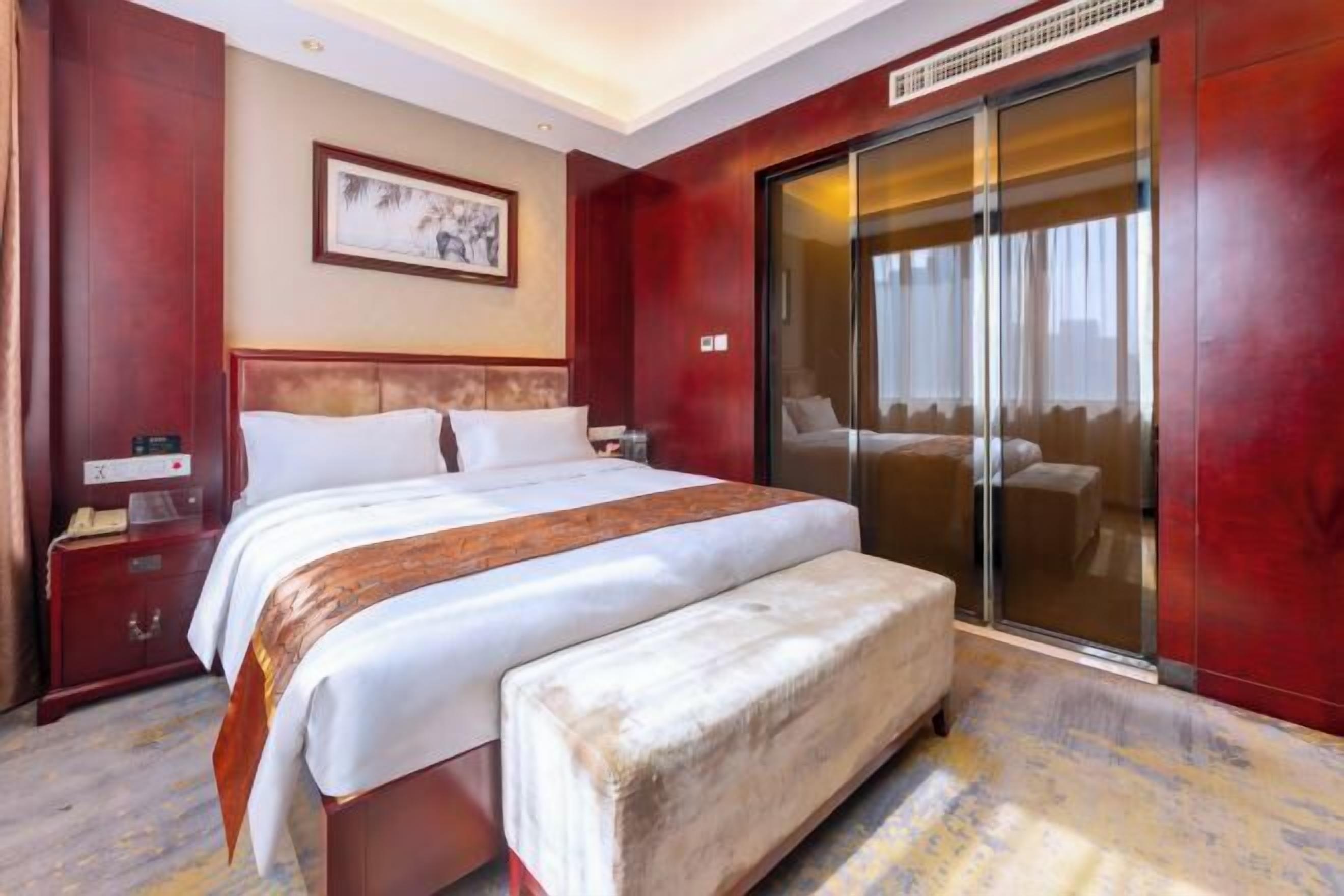 A hotel room with a large bed, a bedside table, a bench, a sliding glass door, and a framed picture on the wall.