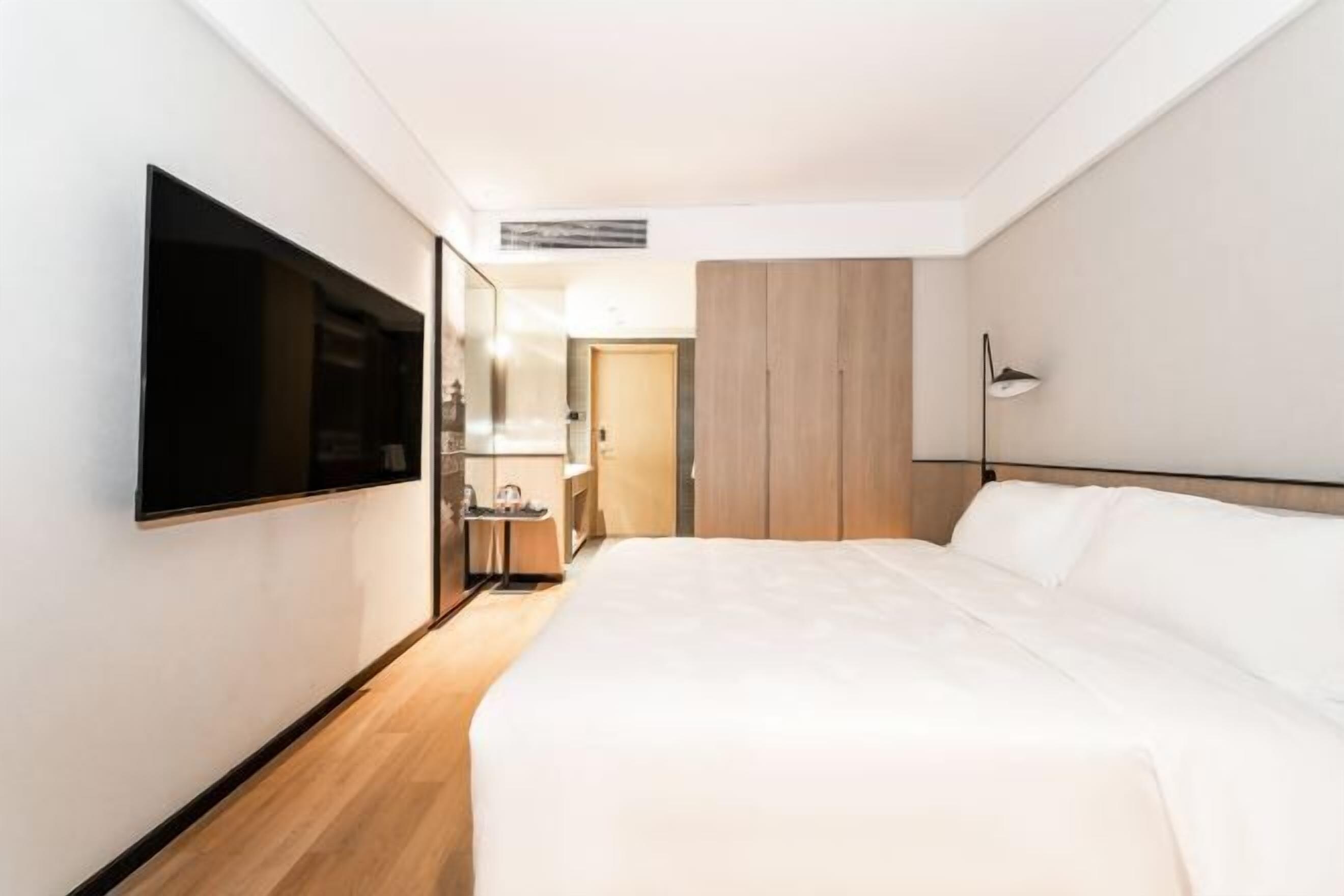 A hotel room with a bed, a TV mounted on the wall, a bedside table, a mirror, and a wardrobe.