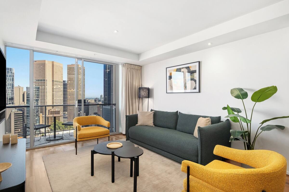 A modern living room with a sofa, two yellow chairs, a coffee table, and a large window offering a city view.