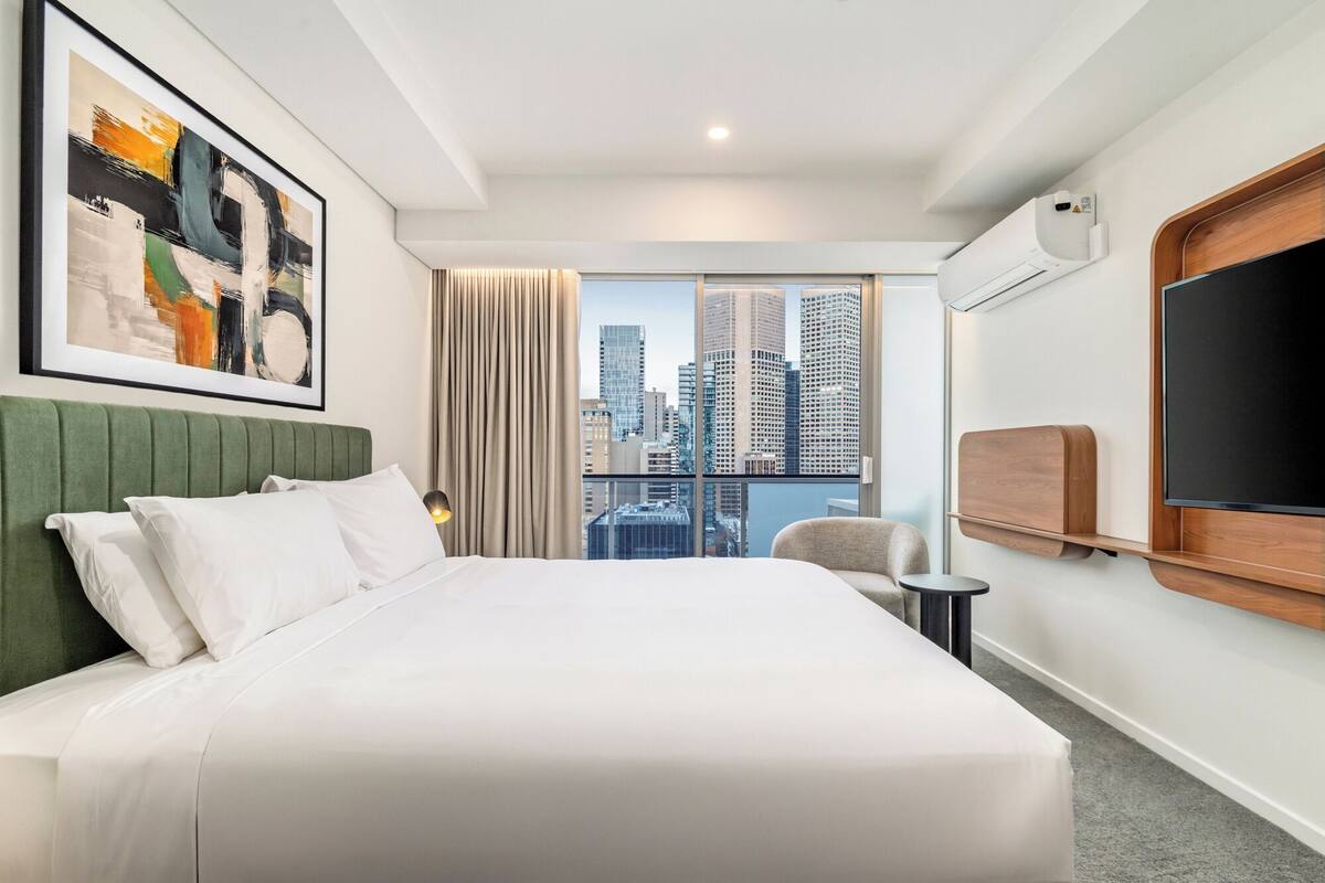A hotel room with a large bed, a city view, a mounted TV, and a framed abstract painting.