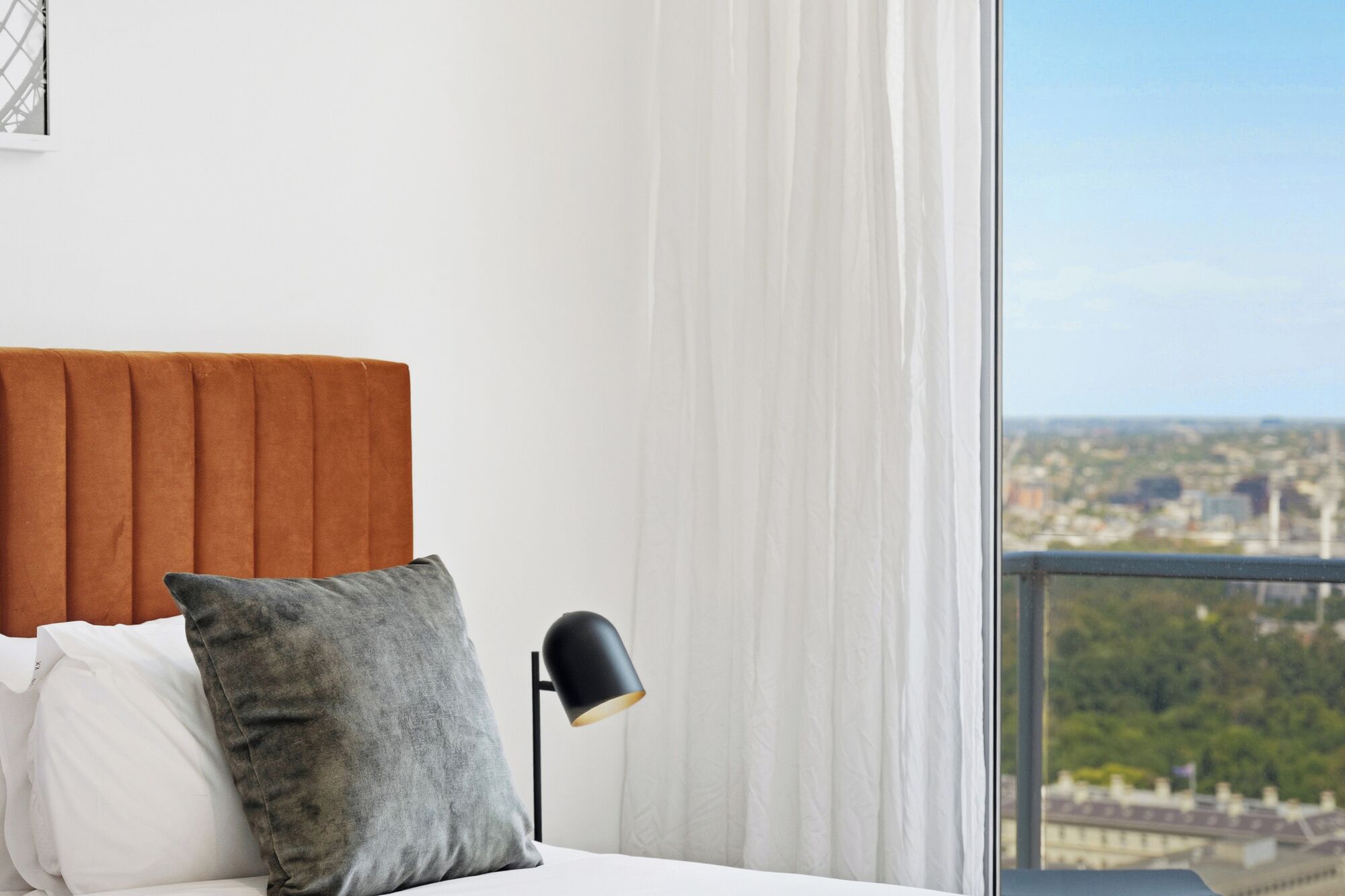 Une chambre à coucher avec un lit, des oreillers, une table de chevet, une lampe et une vue sur le paysage urbain par la fenêtre.
