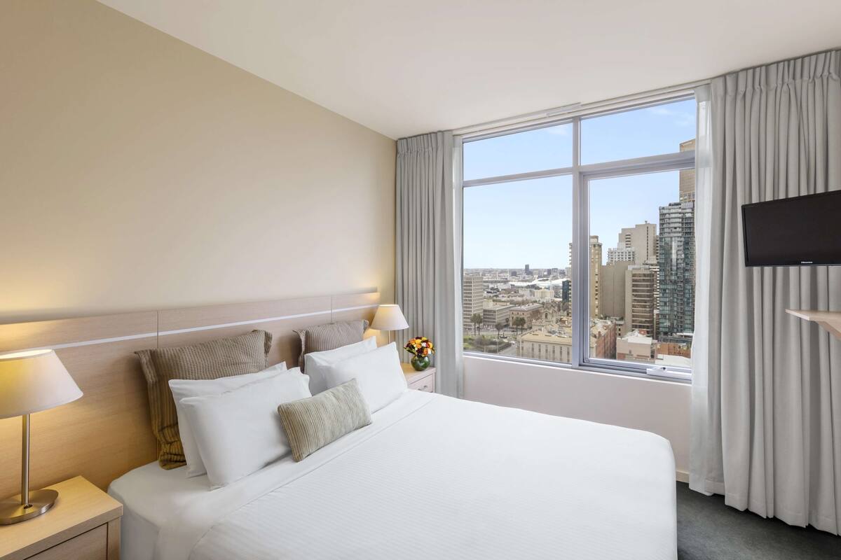 A hotel room with a large bed, a nightstand with a lamp, a TV on the wall, and a window with a city view.
