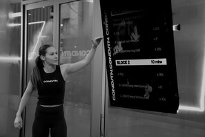 Uma pessoa vestindo top preto e legging com a inscrição 'COND/TNL', em frente a uma placa com instruções de treino.