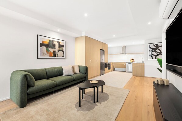 A modern living room with a green sofa, a small coffee table, and a kitchen area with wooden cabinets and appliances.
