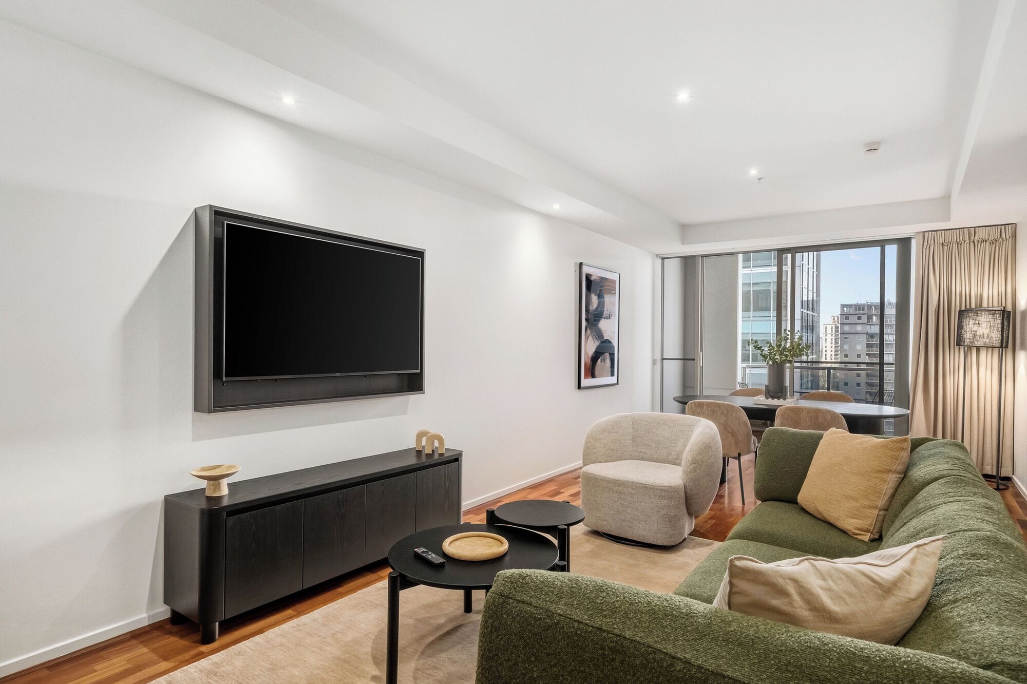 Een moderne woonkamer met een flatscreen televisie, een bank, een eethoek en uitzicht op gebouwen door grote ramen.