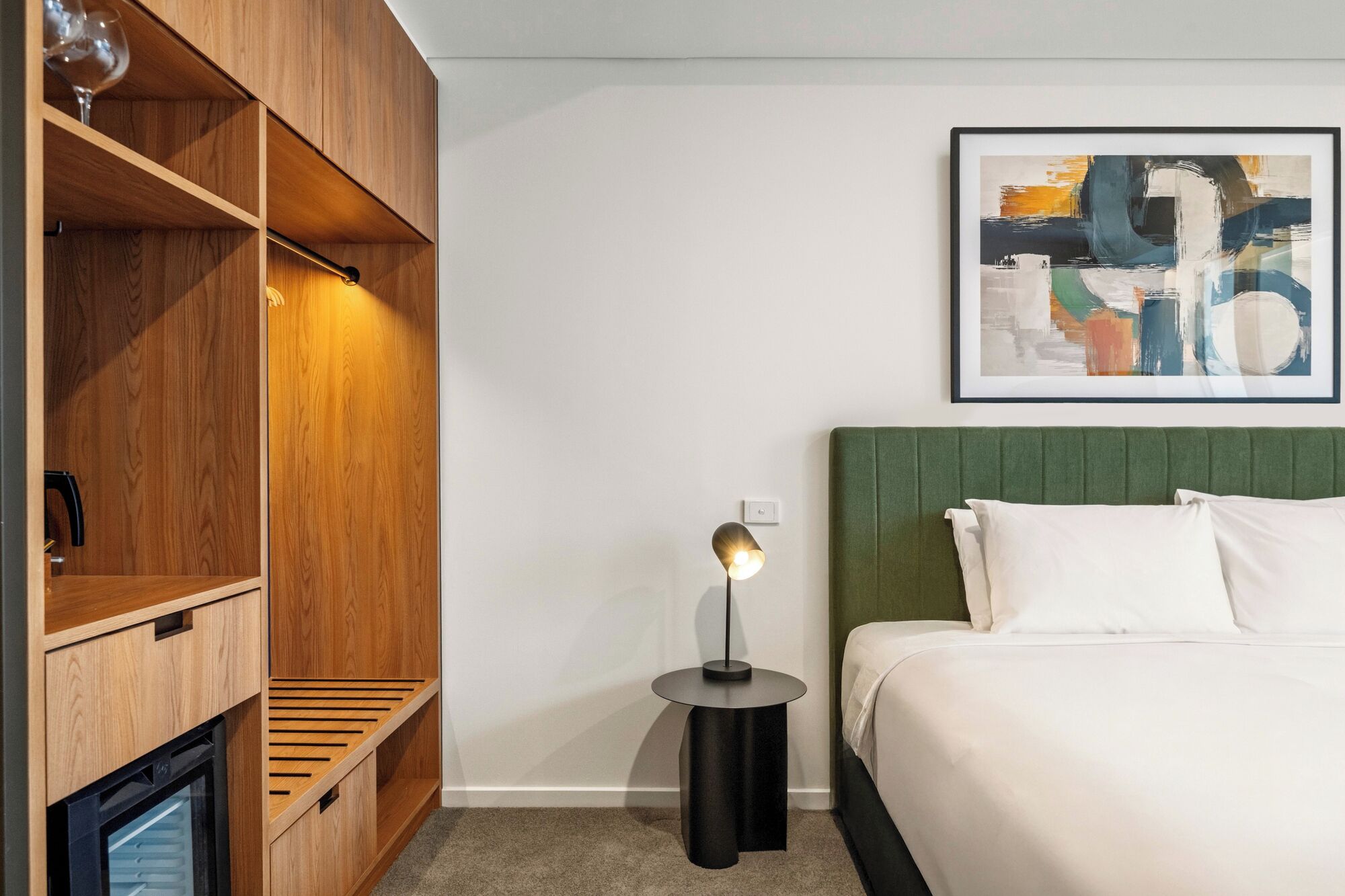A hotel room with a bed, a bedside table with a lamp, a wooden wardrobe, and a framed abstract painting on the wall.