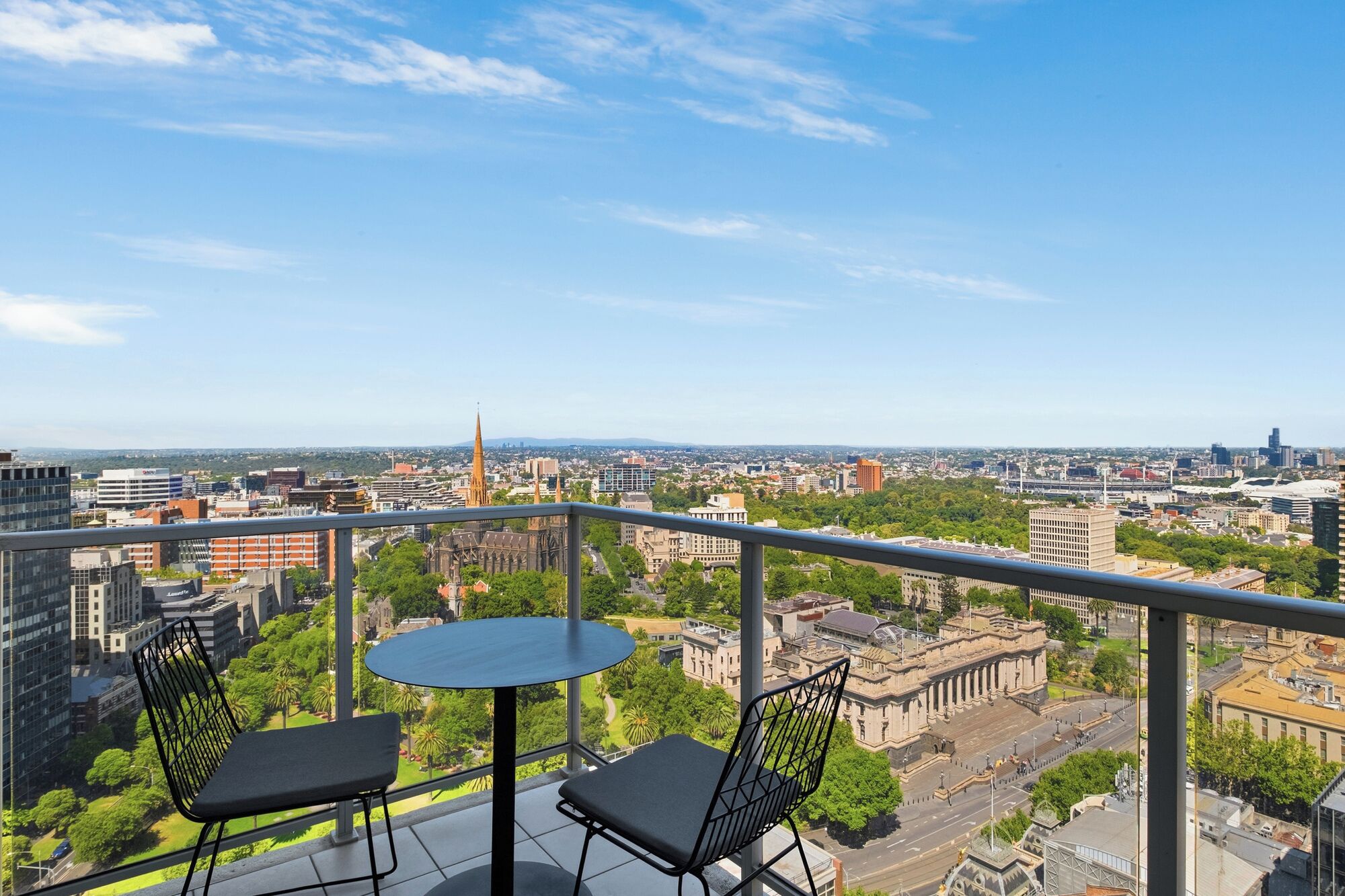 A balcony with two chairs and a table, overlooking a cityscape with buildings and a church spire.