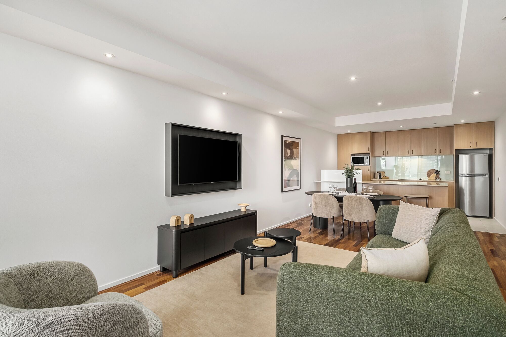 A modern living room with a flat-screen TV, a sofa, a coffee table, and a dining area with a kitchen in the background.