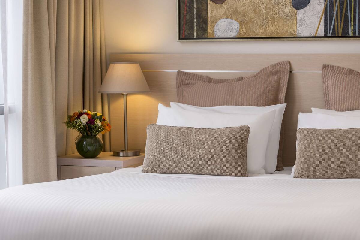 A neatly made bed with white linens and beige pillows, a bedside lamp, and a vase with flowers on a nightstand.