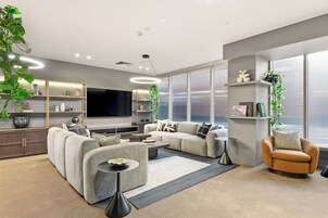 A modern living room with a sofa set, a coffee table, and a TV. There are shelves with decorative items and plants.