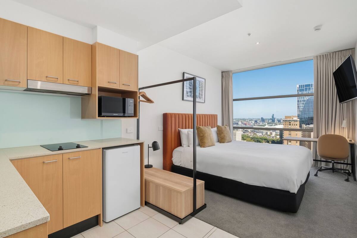 A hotel room with a kitchenette, a bed with a city view, a desk, and a TV mounted on the wall.