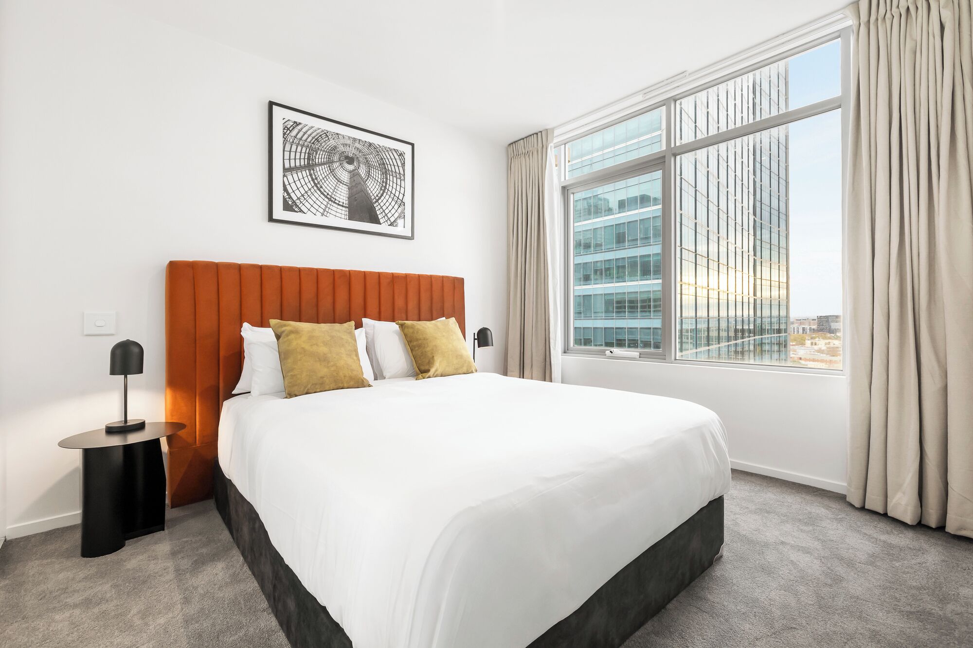 A hotel room with a large bed, a headboard, a framed picture on the wall, a nightstand, and a window with curtains.