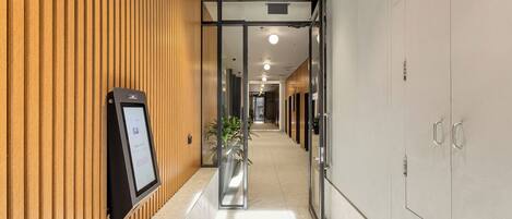 A corridor with wooden paneling on one side, a reception area with a digital display, and a glass door leading to another room.