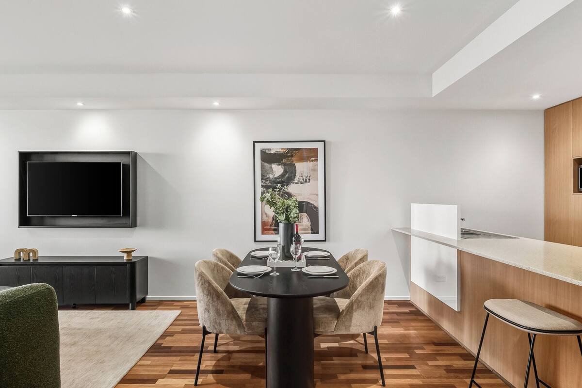 A modern kitchen with a dining area, a flat-screen TV, and a framed artwork above the table.