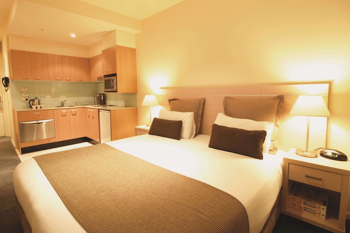 Una habitación de hotel con cama, lámparas de noche, mesita de noche, un escritorio pequeño y una kitchenette.
