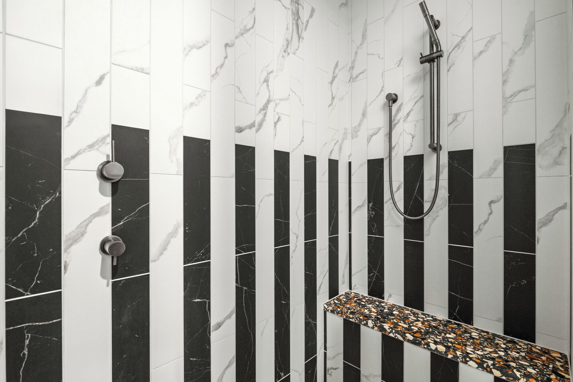 A bathroom with black and white marble tiles, a shower with a handheld showerhead, and a small bench with a terrazzo top.