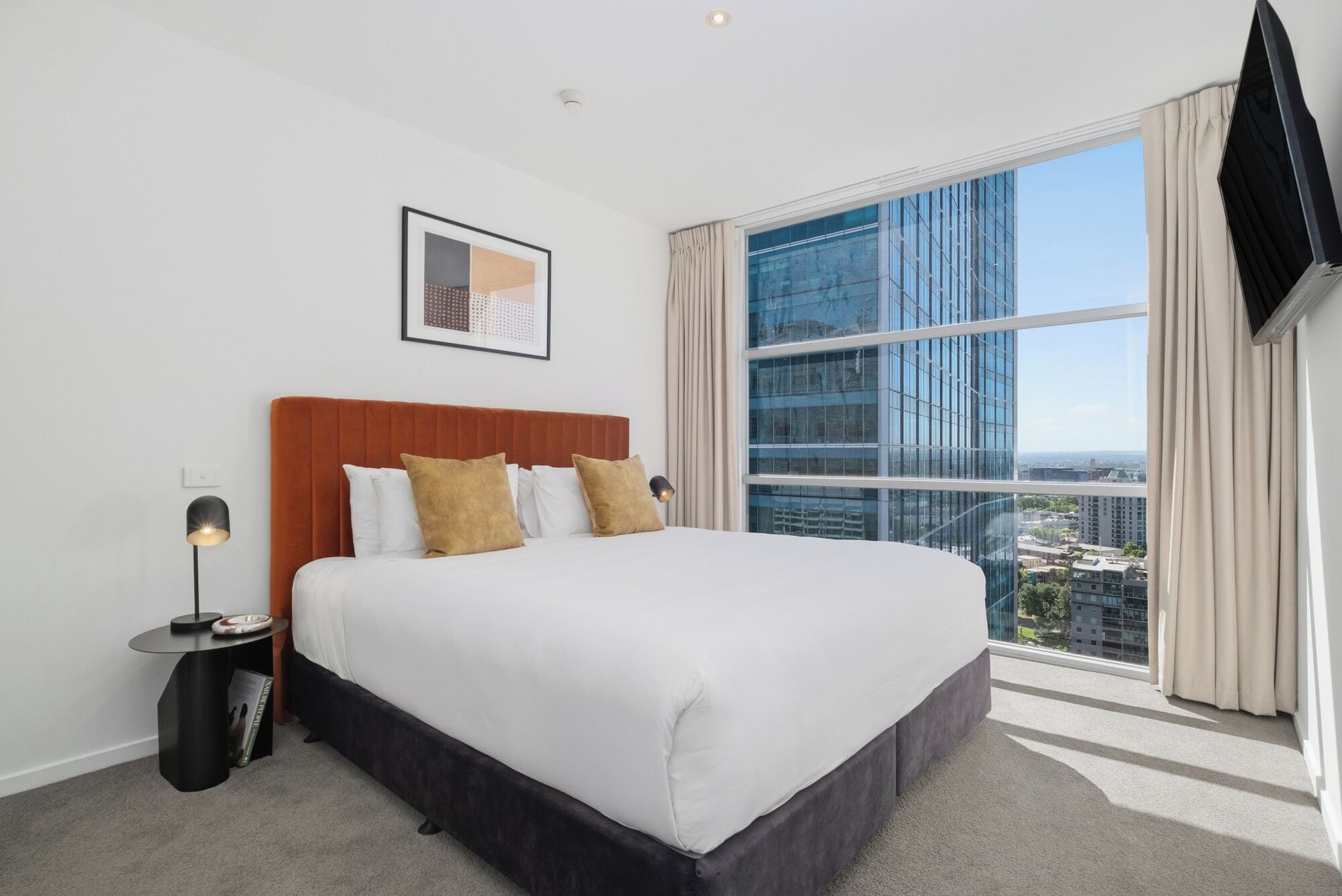 Habitación de hotel con una cama grande, vistas de la ciudad a través de ventanas del suelo al techo, un televisor de pantalla plana montado y una obra de arte abstracta enmarcada en la pared.